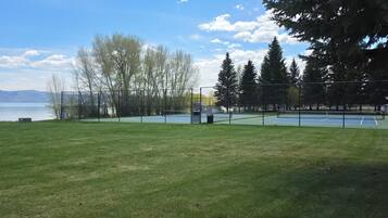 A tennis court with a net, surrounded by a fence, and a body of water in the background.
