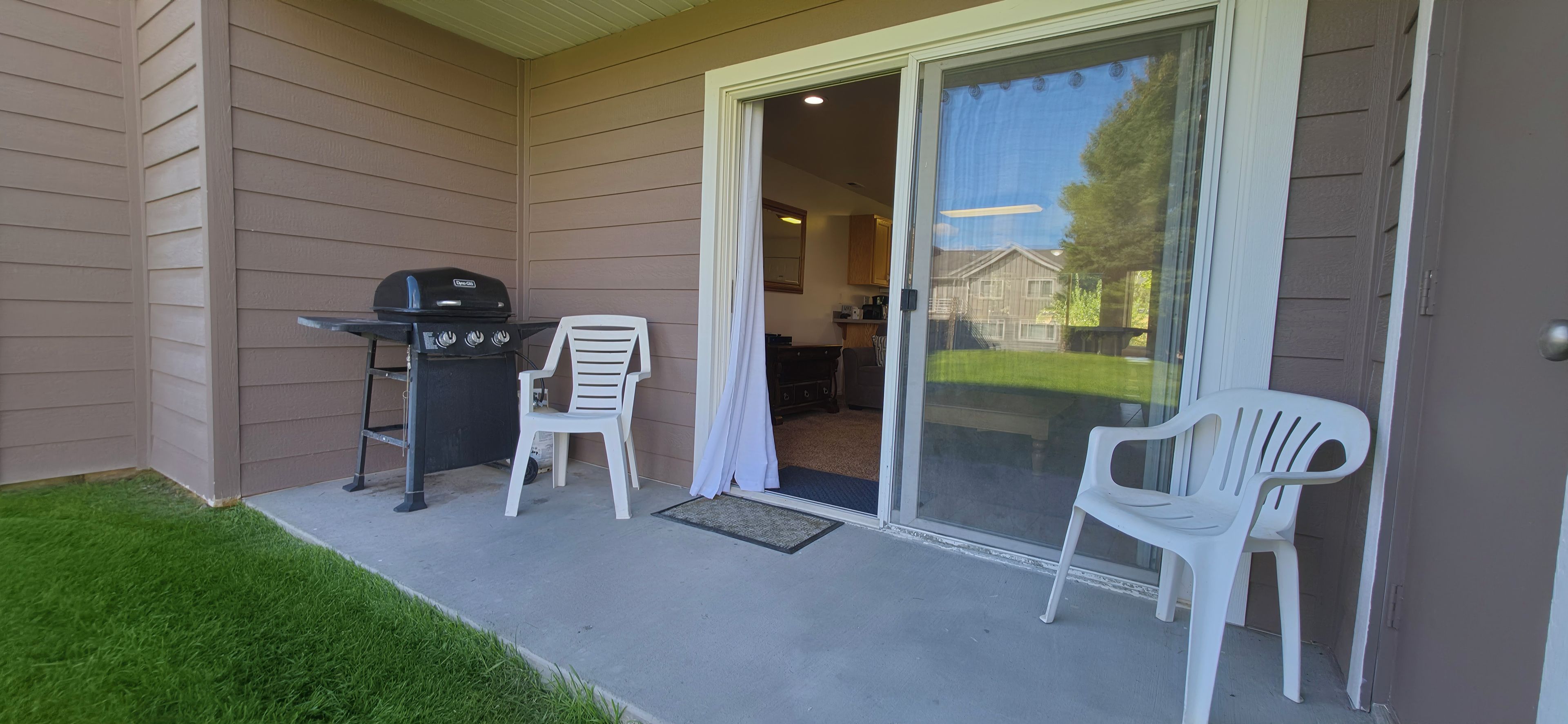 A patio with a grill, two white chairs, and a sliding glass door leading to a backyard.