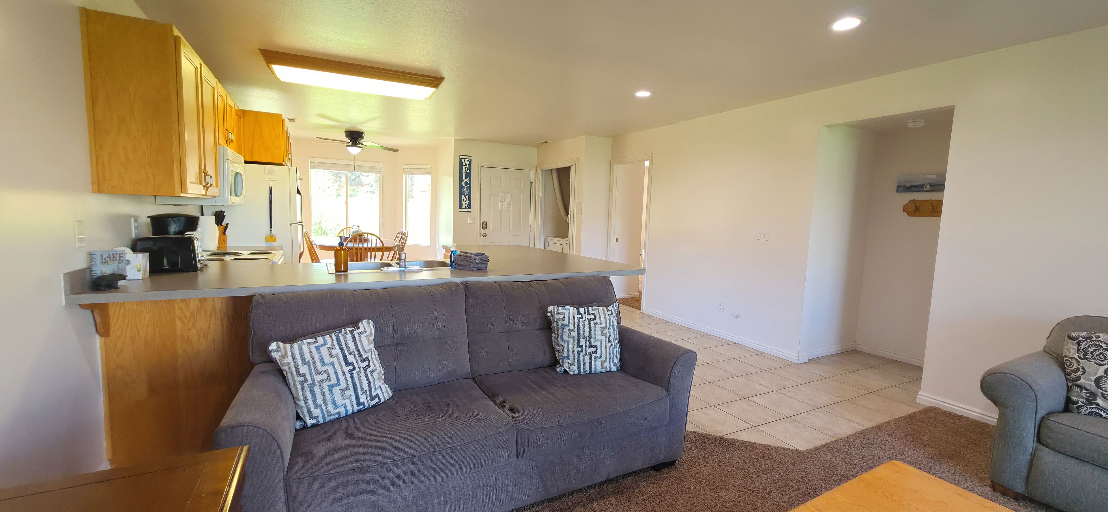 A modern kitchen with wooden cabinets, a grey sofa with patterned cushions, and a dining area with a table and chairs.
