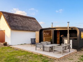 Ein Gebäude mit einem Strohdach, wobei ein offener Bereich einen Kamin, Sitzgelegenheiten im Freien und eine Holzterrasse zeigt.