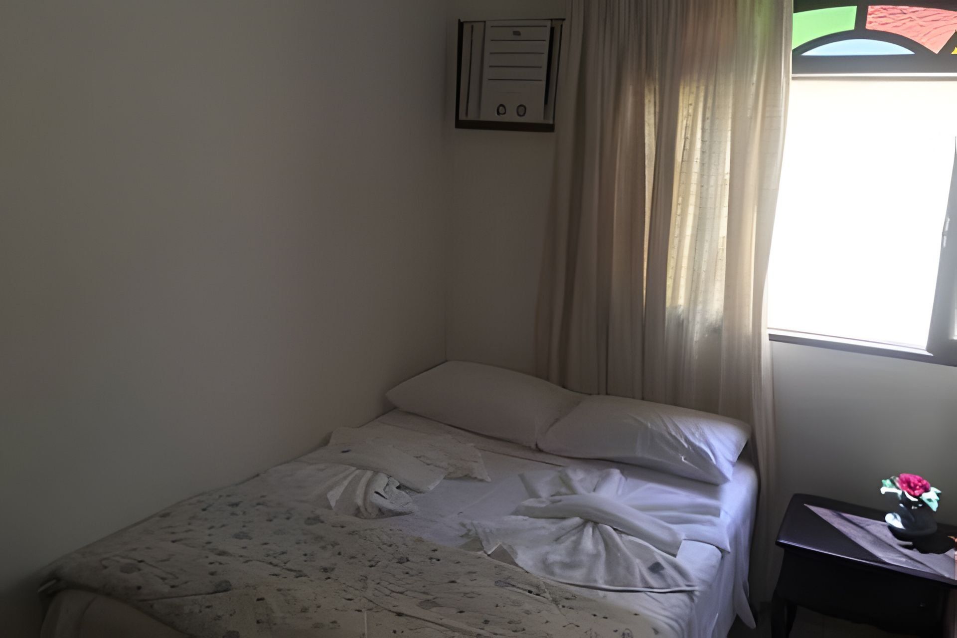 Una cama individual con cubrecama y almohadas blancas, una mesita de noche con un florero y una ventana con cortinas.