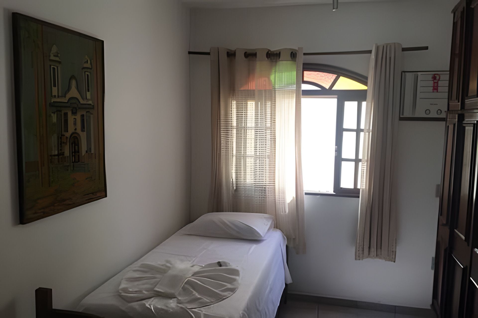 A single bed with a white sheet and a decorative pillow, a window with sheer curtains, a wall-mounted thermostat, and a framed picture on the wall.