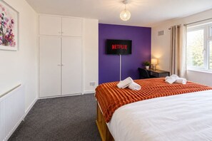 Ein Hotelzimmer mit einem Bett, einem an einer lilafarbenen Wand montierten Fernseher, einem Schreibtisch mit Stuhl und einem Fenster mit Vorhängen.