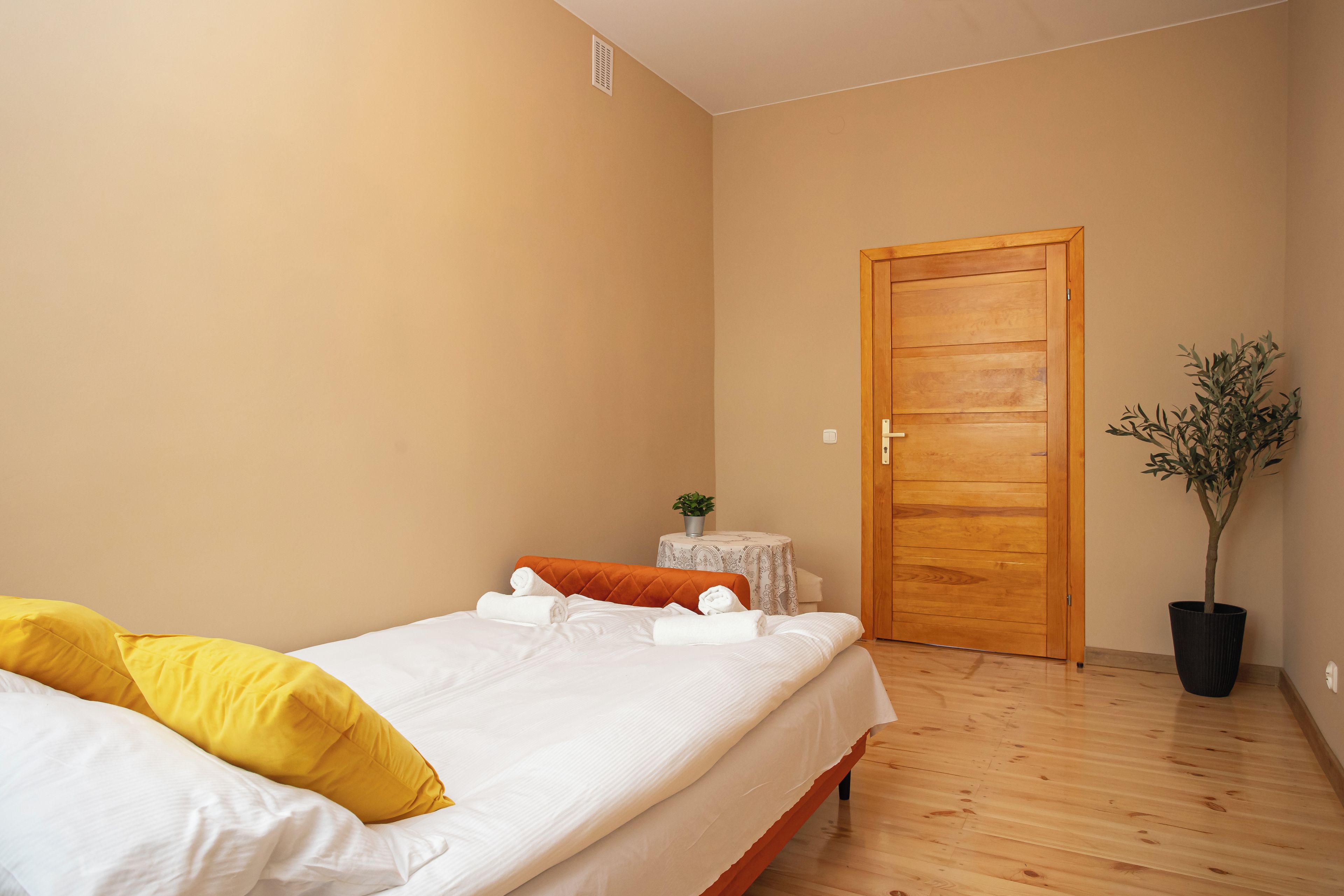 A bedroom with a bed, a wooden door, a small table with a plant, and a potted plant beside the door.