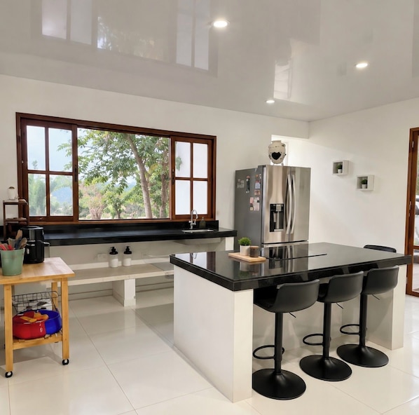 Una cocina moderna con una isla central, taburetes de bar, un refrigerador y un ventanal con vistas a árboles.