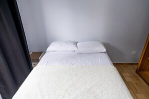 Un lit avec une literie blanche dans une pièce dont le sol est en bois et dont un des murs est uni.