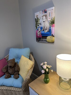 A room with a wicker chair, a teddy bear, a vase with flowers, a lamp, and a painting on the wall.