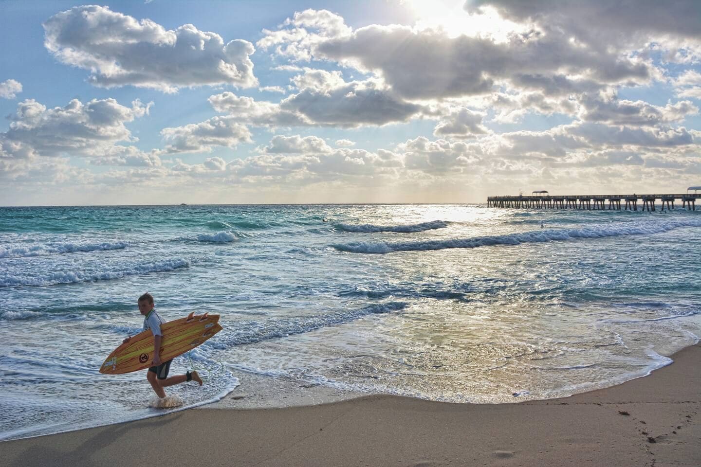 Une plage avec des vagues, une jetée en arrière-plan et une personne qui porte une planche de surf.