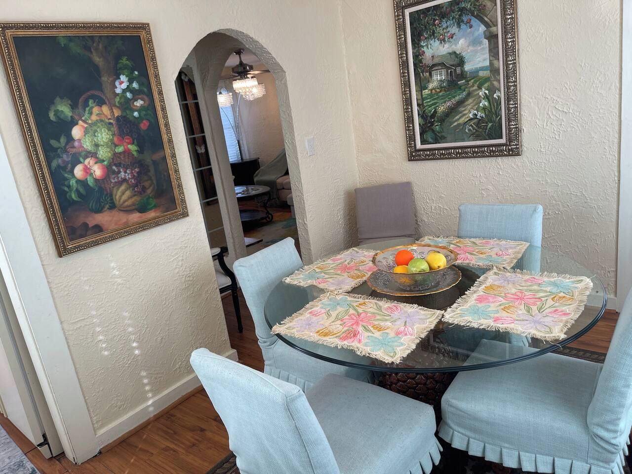 Un coin repas avec une table ronde en verre, des chaises à revêtement bleu clair et des sets de table à motifs floraux.