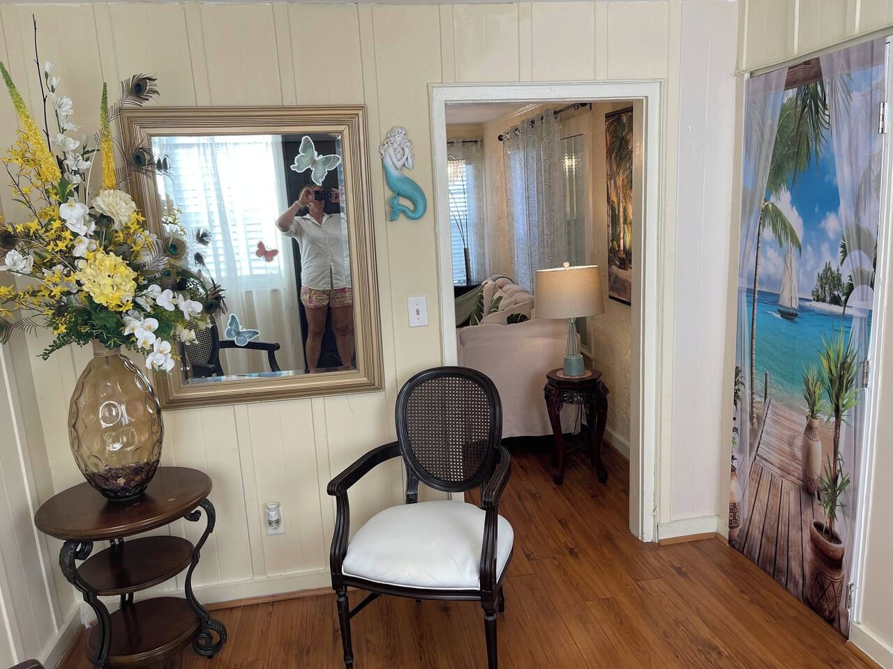 Une pièce avec un plancher en bois, une chaise, un miroir, un vase de fleurs et une porte qui donne sur une autre pièce.