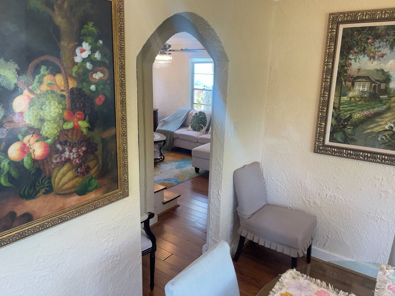 Une pièce avec un fauteuil blanc, un tableau et, à travers une arche, une vue sur une autre pièce où se trouvent un canapé et une fenêtre.