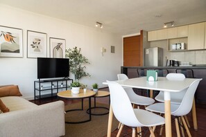 Een moderne woonkamer met een flatscreen televisie, een bank, een eettafel met stoelen en een keuken met een magnetron en een koelkast.