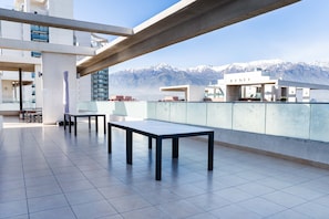 A rooftop terrace with a view of snow-capped mountains, featuring a long table and chairs, and a glass railing. - Kennedy Apartments by Andes with Parking (Santiago)