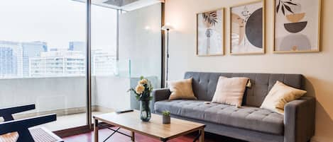 A modern living room with a grey sofa, a wooden coffee table, and a city view through large windows.