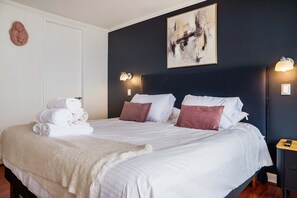 A neatly made bed with white linens and a folded towel, a dark wall with a painting, and two wall-mounted light fixtures. - Kennedy Apartments by Andes with Parking (Santiago)