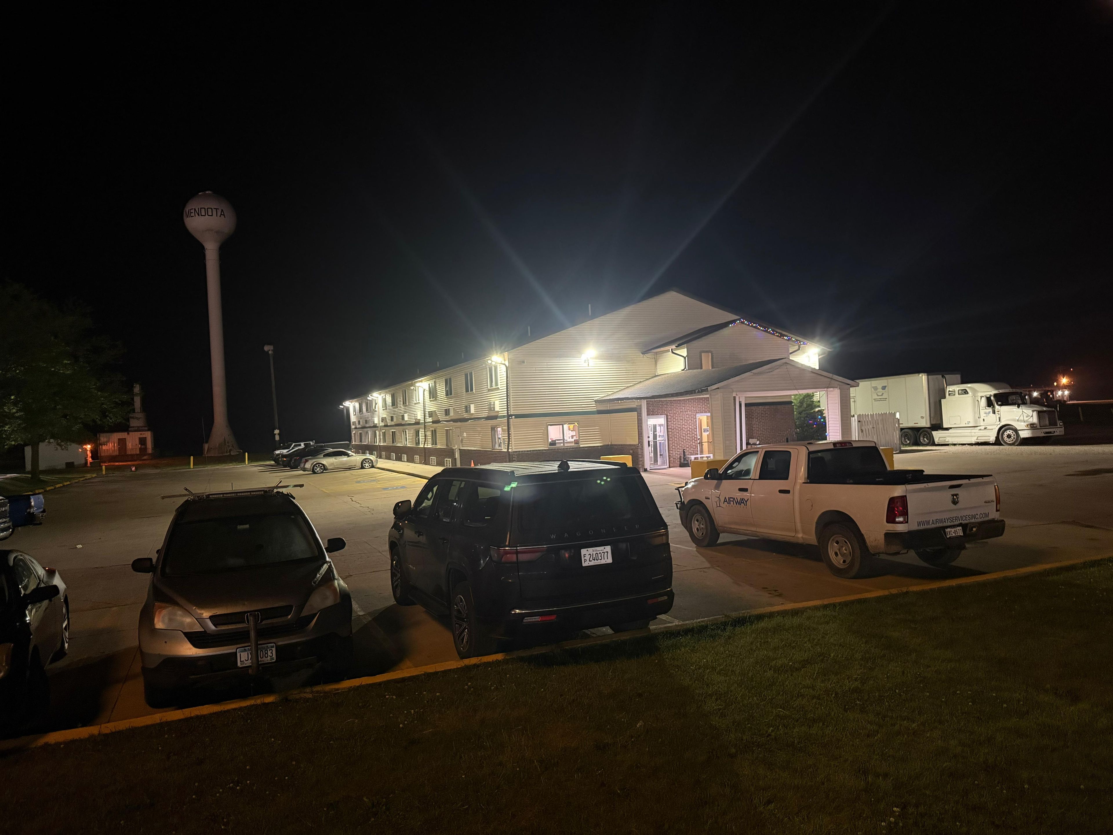 Un parking avec plusieurs véhicules, un bâtiment doté d’une enseigne lumineuse et une grande structure en arrière-plan.
