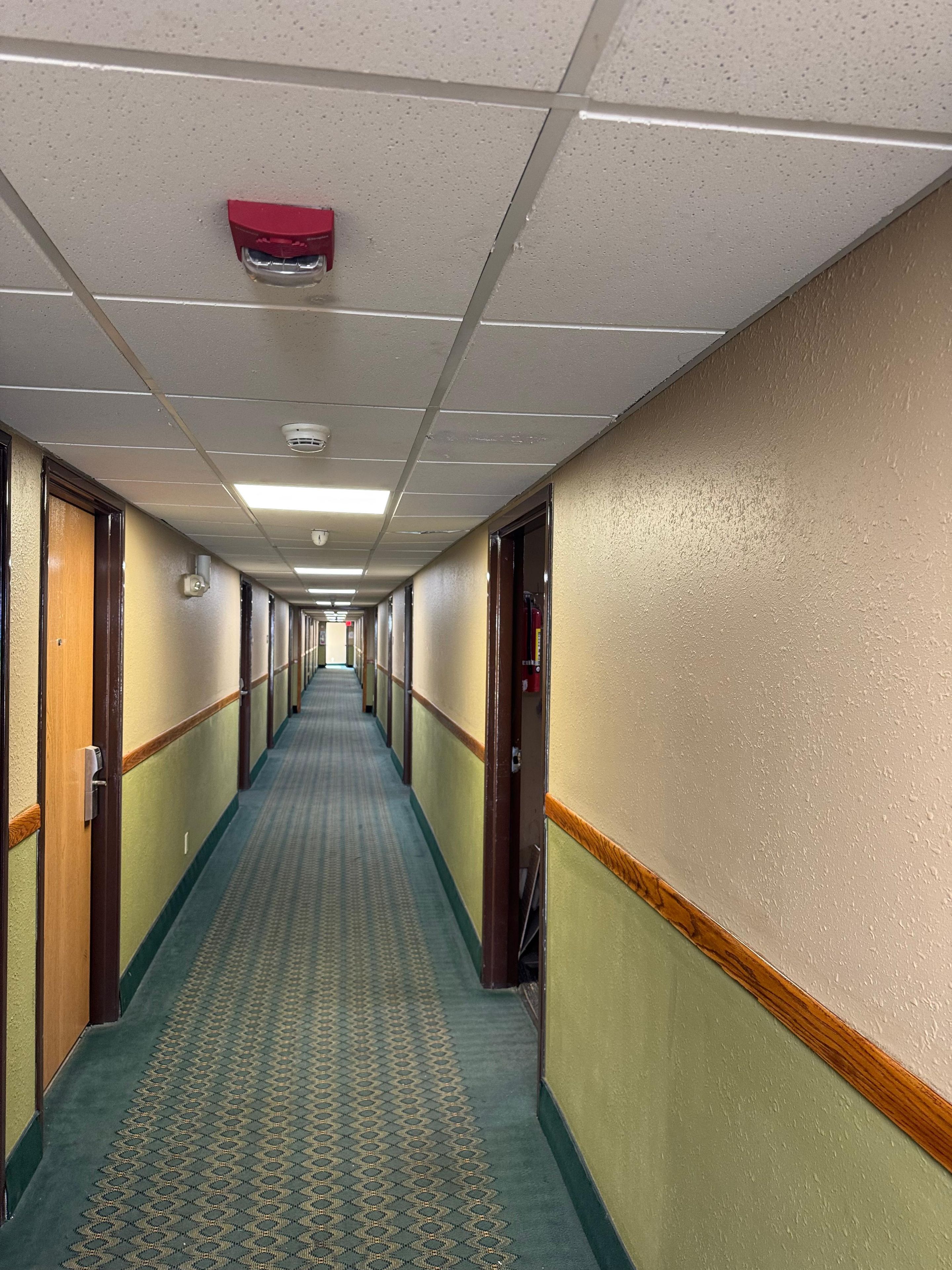 En lång, smal hotellkorridor med ett mönstrat golv, dörrar på båda sidor och en brandlarm på taket.