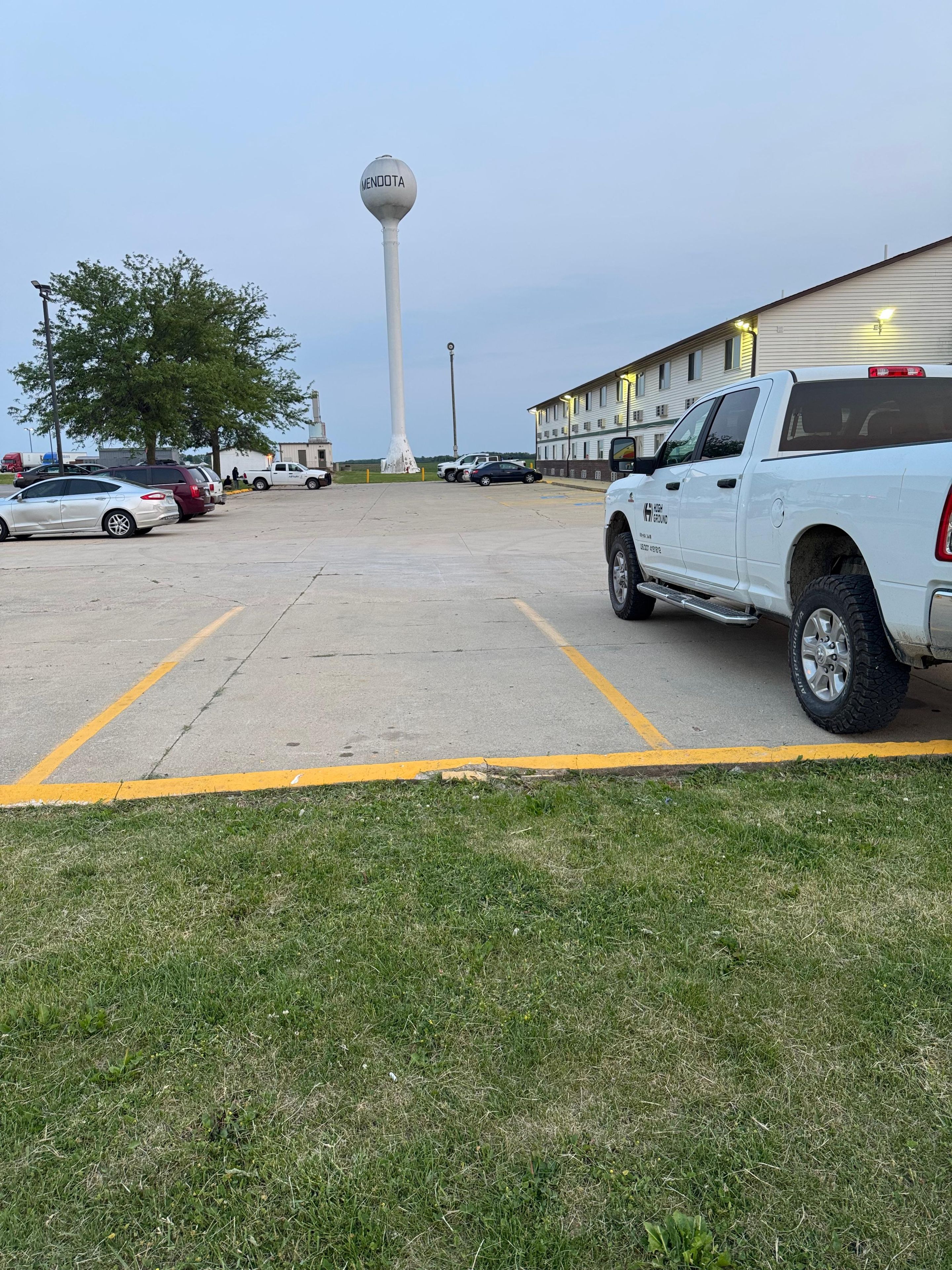 A parking lot with a white pickup truck, a white building, and a tall structure in the background.