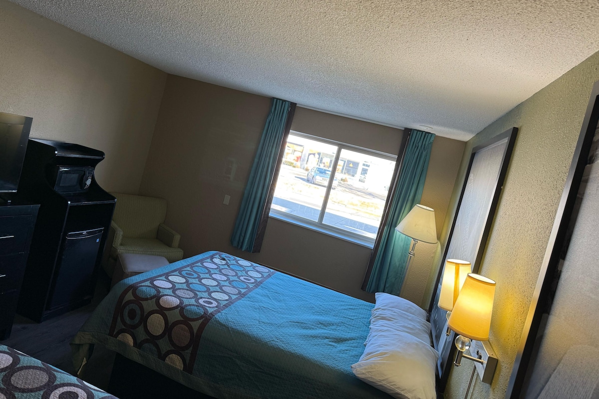 A hotel room with a bed, a desk with a computer, a window with curtains, and a lamp.