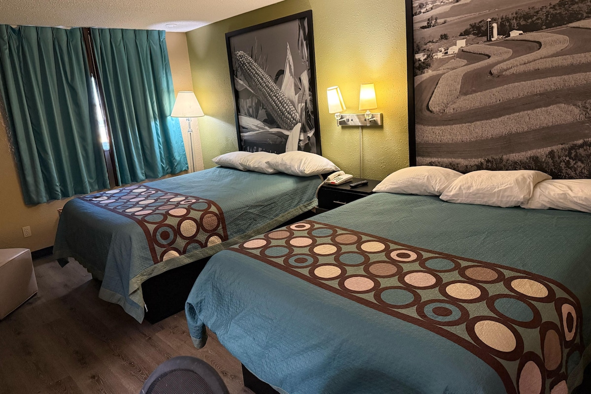 A hotel room with two beds, a lamp, a telephone, and a framed landscape painting.