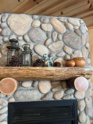 Una chimenea de piedra con un estante de madera encima, donde se exhiben faroles, piñas y cerámica.