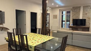 Un comedor con mesa de vidrio y sillas de madera, una sala de estar con sofá y televisión, y una pared de piedra.