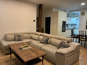 Ein Wohnzimmer mit Ecksofa, Couchtisch aus Holz und einer Küche im Hintergrund.