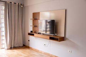Ein modernes Wohnzimmer mit einem Flachbildfernseher an einer Holzablage, einem Fenster mit Vorhängen und einem Fliesenboden.