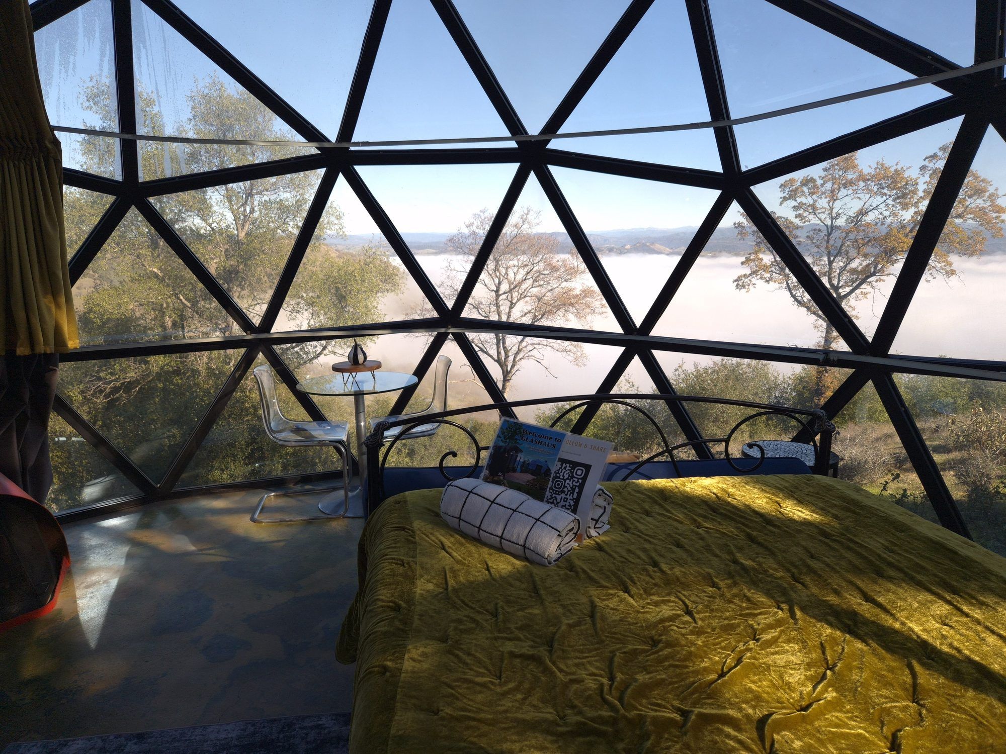 A room with a bed, a table, and a chair, with a view of trees and a body of water through a geometric window structure.