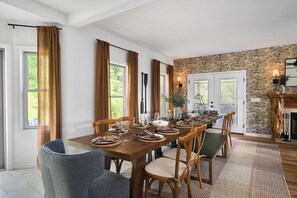 Ein Esszimmer mit einem langen Holztisch, gedeckt für sechs Personen, mit großen, mit Vorhängen drapierten Fenstern, einer Steinwand und einem Kamin mit Kaminfeuer.