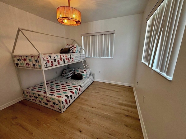 Une pièce avec un lit superposé, un luminaire suspendu, une fenêtre avec des stores et un plancher en bois.