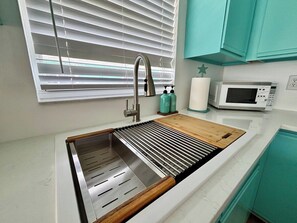 Een keuken met een roestvrijstalen spoelbak, een houten snijplank, een magnetron en kastjes in een blauwgroene kleur.