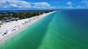 Une plage avec une longue étendue de sable, une eau turquoise et limpide, et quelques bâtiments visibles au loin.