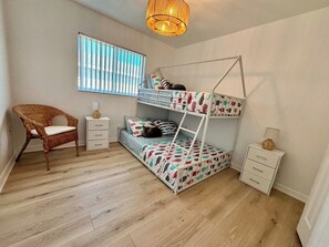 Een kinderbed met twee stapelbedden, een bed met een patroon, een rieten stoel, een nachtkastje en een raam met jaloezieën.