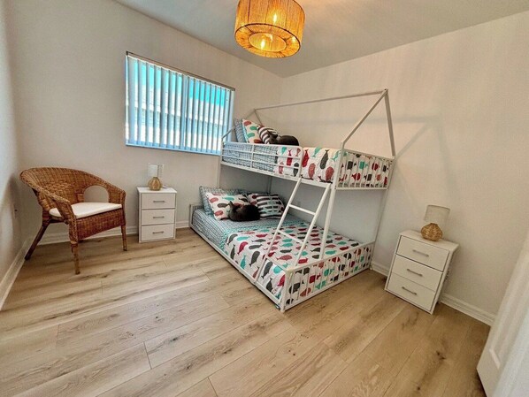 Un lit superposé pour enfant, avec une couvre-lit à motifs, une chaise en osier, une table de chevet et une fenêtre avec des stores.
