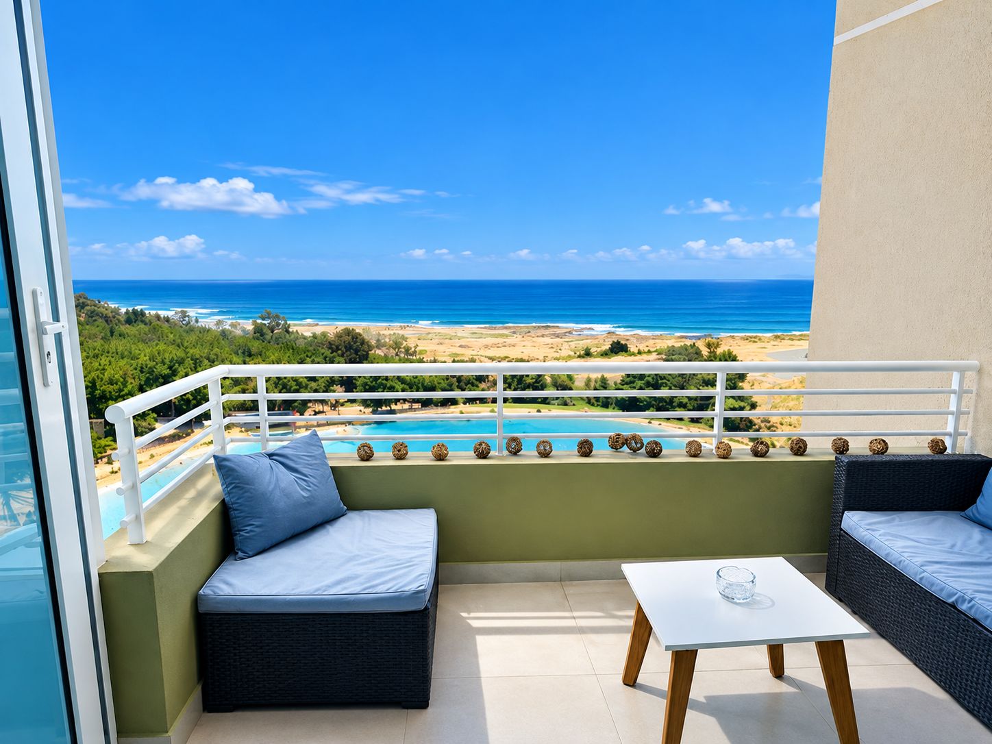 Ein Balkon mit Blick auf Strand und Meer, ein weißer Tisch und zwei Sitzbereiche mit Polstern.