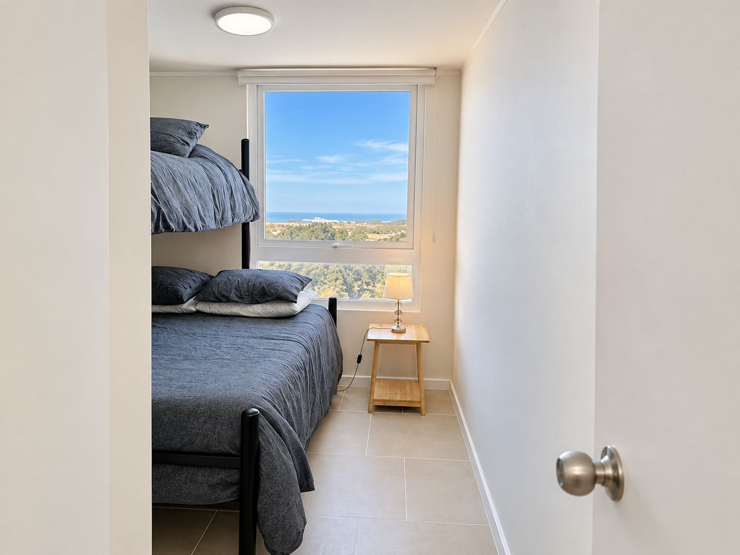 A bedroom with a bunk bed, a bedside table with a lamp, and a view of the landscape through a window.
