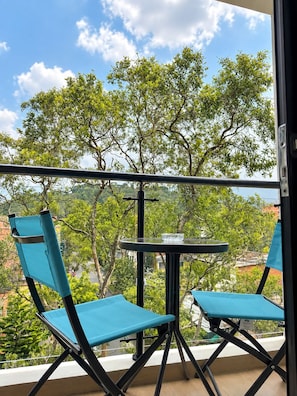 Ein Balkon mit zwei blauen Stühlen und einem kleinen Tisch, mit Blick auf eine üppige grüne Landschaft mit Bäumen und Gebäuden im Hintergrund.