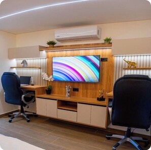 Un espacio de oficina moderno con una gran televisión de pantalla plana montada en una pared de madera, dos sillas de oficina, un escritorio con una computadora portátil y una unidad de aire acondicionado sobre la televisión.