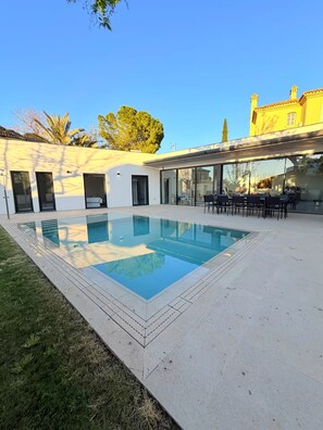 Un espace piscine extérieur moderne, avec un ciel dégagé, un coin repas avec des tables et des chaises, et un bâtiment doté de grandes fenêtres et d’une façade jaune.
