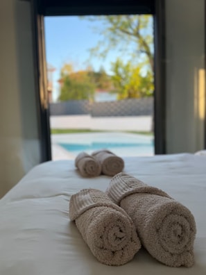 Ein Bett mit zusammengerollten Handtüchern und Blick auf einen Swimmingpool durch ein Fenster.