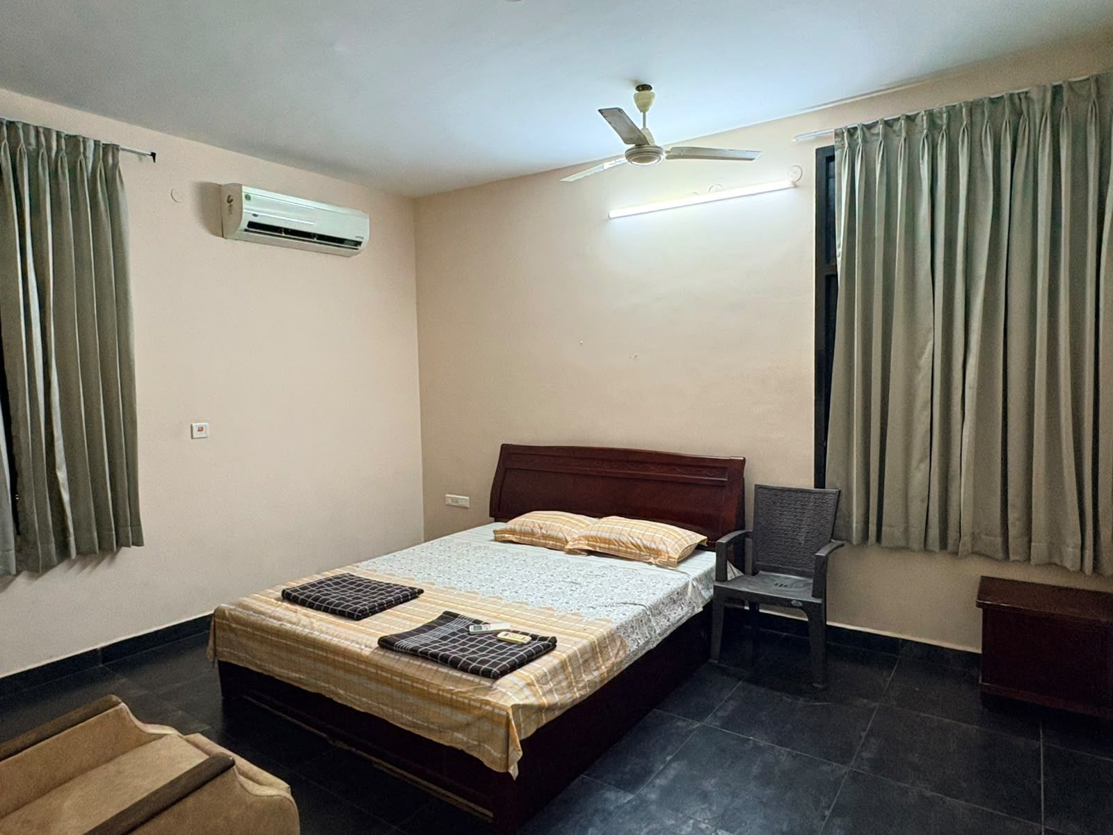 Een slaapkamer met een bed, een airconditioner, een plafondventilator, een stoel en gordijnen.