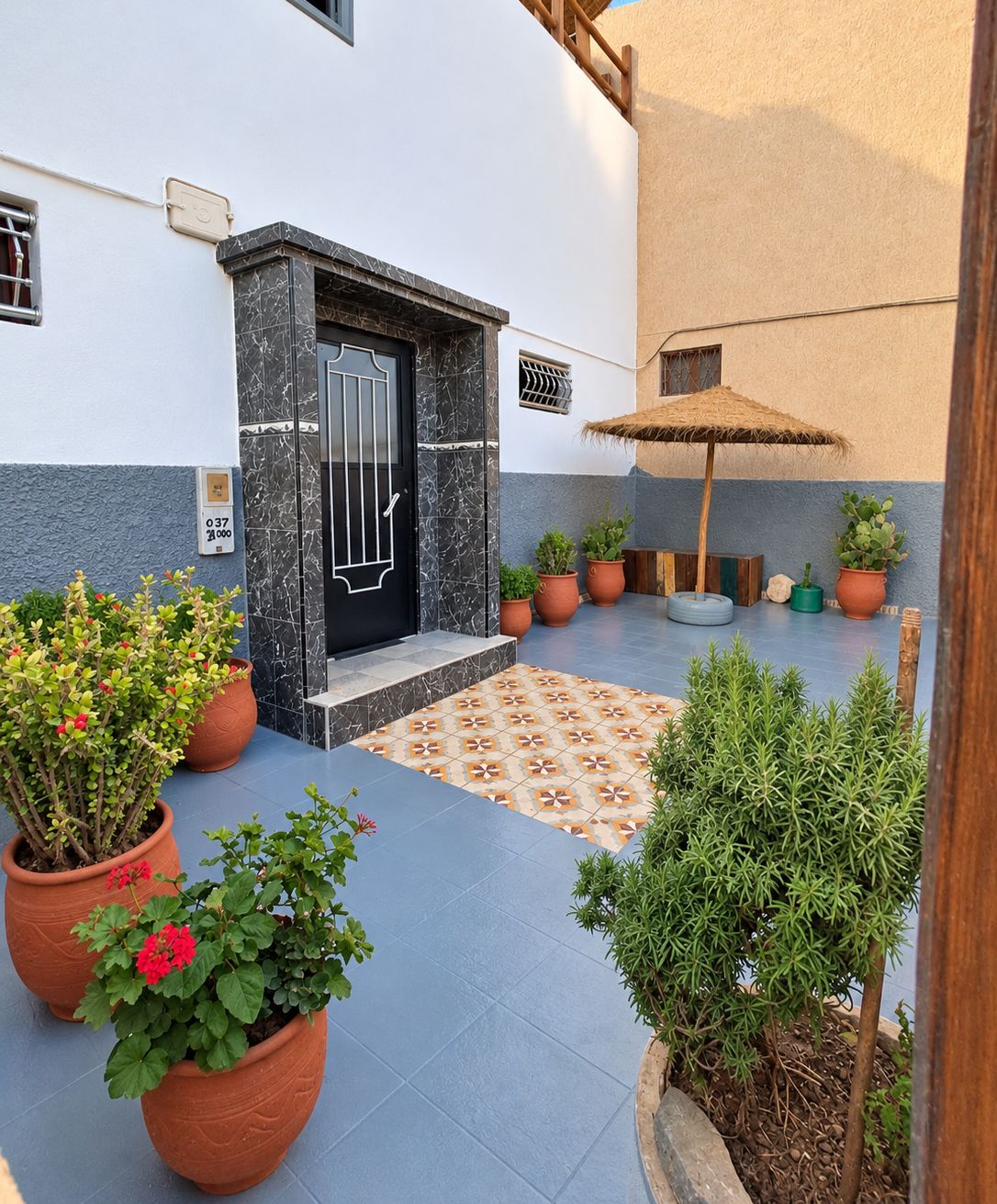 A courtyard with a tiled floor, potted plants, a door with a metal gate, and a small umbrella.