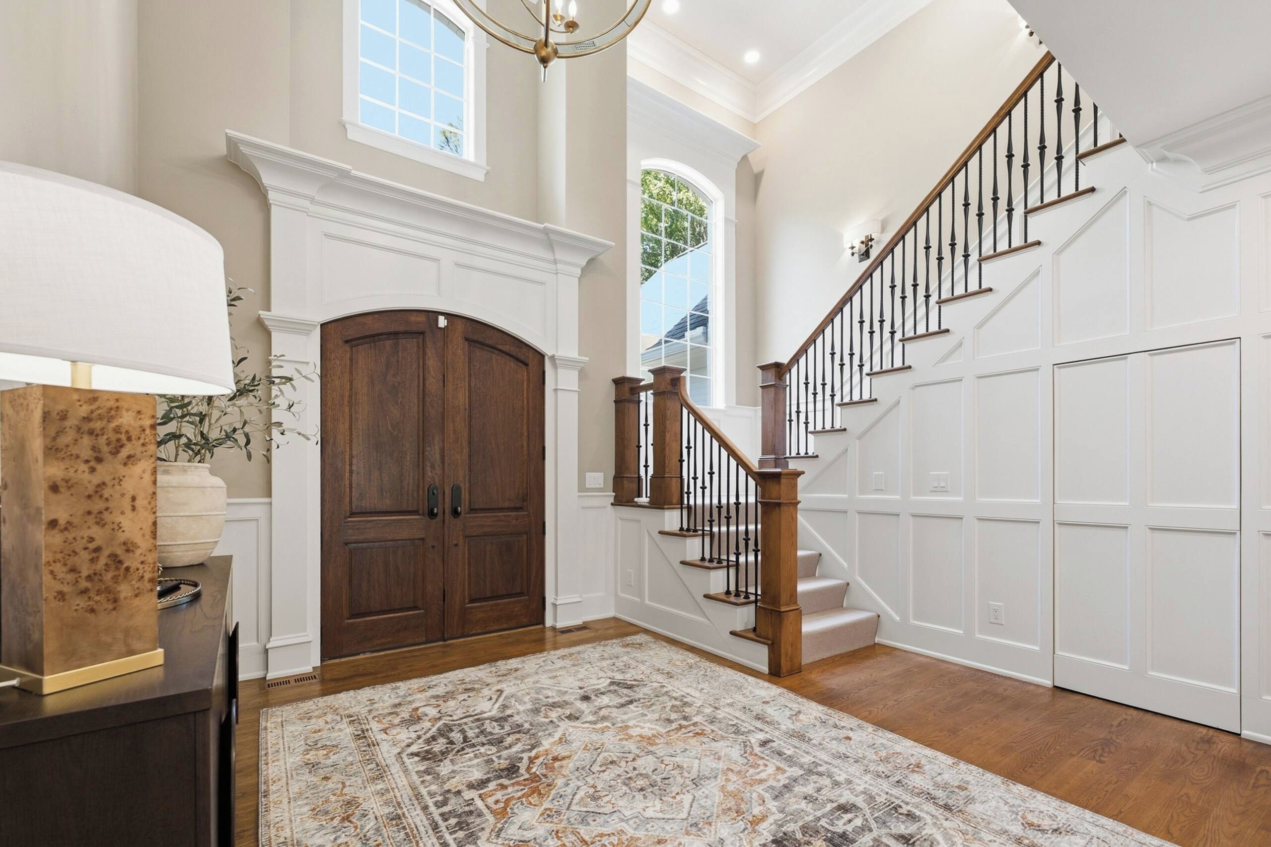 A spacious entryway with a grand wooden staircase, a large double door, and a patterned rug.