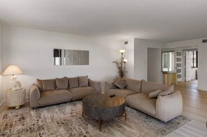 Un salon moderne avec un canapé d’angle beige, une table basse ronde et un tapis à motifs. On y trouve une lampe sur une table d’appoint et une plante décorative.