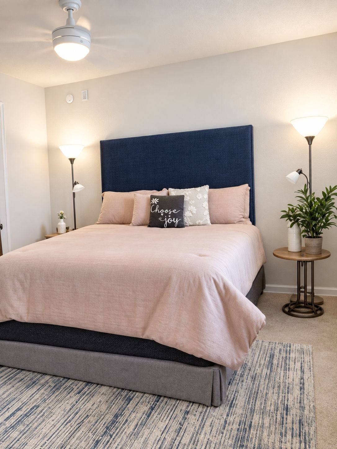 A neatly arranged bedroom with a bed, bedside lamps, a ceiling fan, and a small table with a plant.