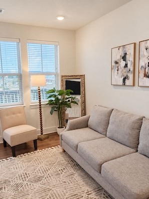 En stue med en beige sofa, en stol, en gulvlampe, en potteplante og to abstrakte malerier på væggen.