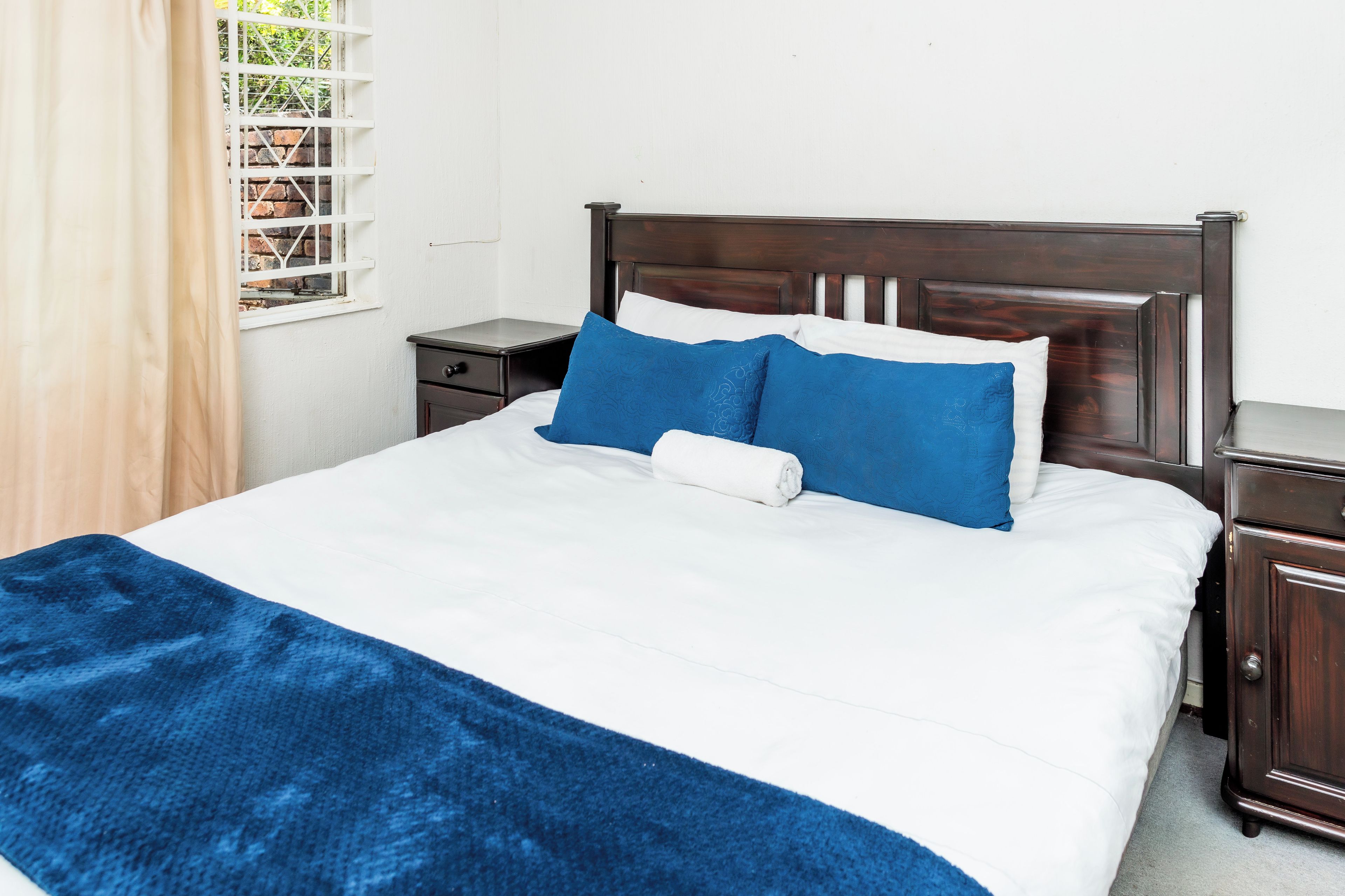 A bed with white bedding and blue pillows, a wooden headboard, a nightstand, and a window with a view of greenery.
