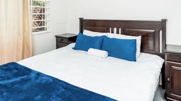 A bed with white bedding and blue pillows, a wooden headboard, a nightstand, and a window with a view of greenery.