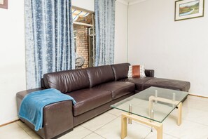 A brown leather sofa with a glass coffee table in a living room with a view of a brick wall and blue curtains.